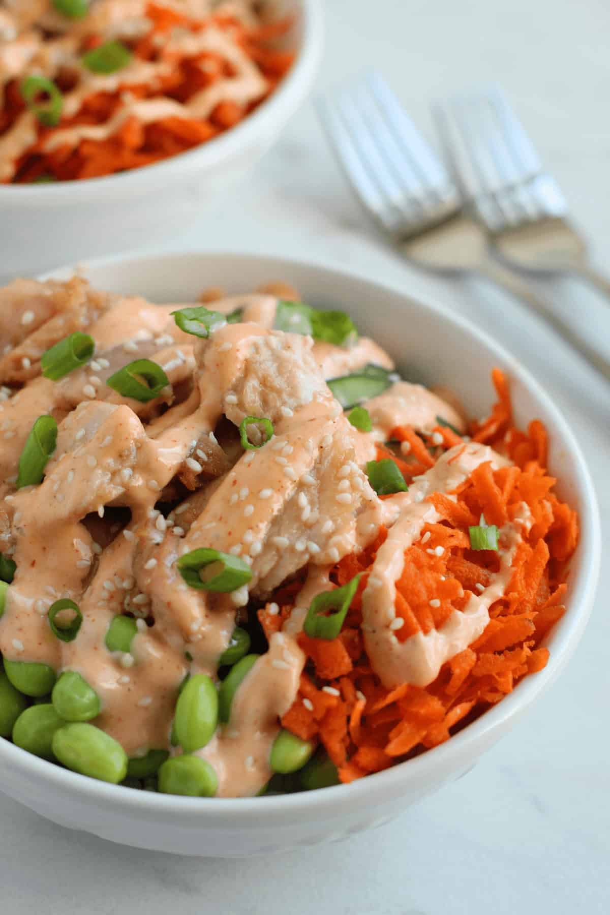 A bowl of mixed salad with shredded carrots, edamame, diced chicken, sesame seeds, chopped green onions, and creamy dressing sits invitingly next to a tempting teriyaki salmon bowl. In the background, a second bowl and two forks await your culinary delight.