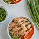 Two bowls of salad with chicken, edamame, shredded carrots, and creamy dressing sit invitingly. Nearby are green onions and a small bowl of fresh edamame on the marble surface, reminiscent of vibrant teriyaki salmon bowls that tantalize the senses.