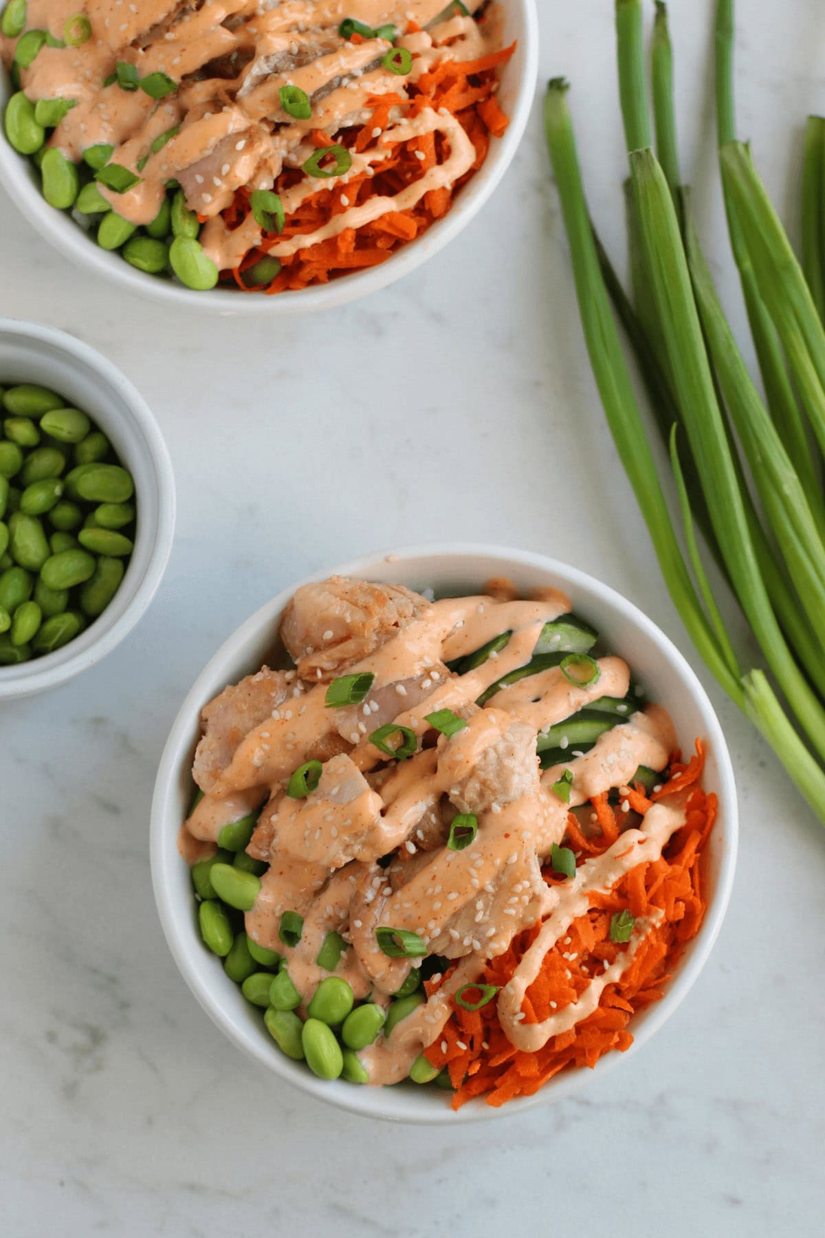 Two bowls of salad with chicken, edamame, shredded carrots, and creamy dressing sit invitingly. Nearby are green onions and a small bowl of fresh edamame on the marble surface, reminiscent of vibrant teriyaki salmon bowls that tantalize the senses.