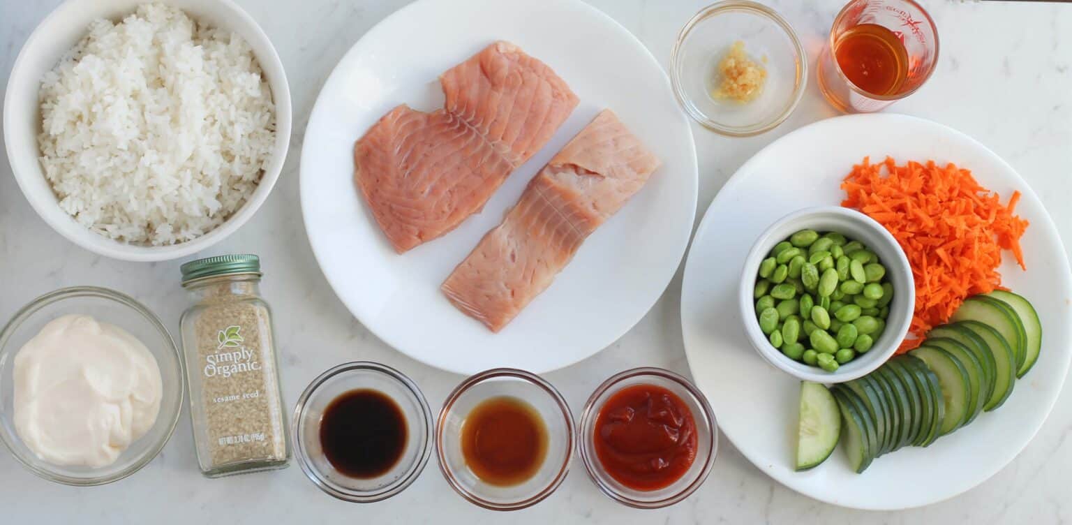 Ingredients fish include cooked rice, two raw fish fillets, grated carrots, edamame, cucumber slices, and various condiments in small bowls on a countertop.