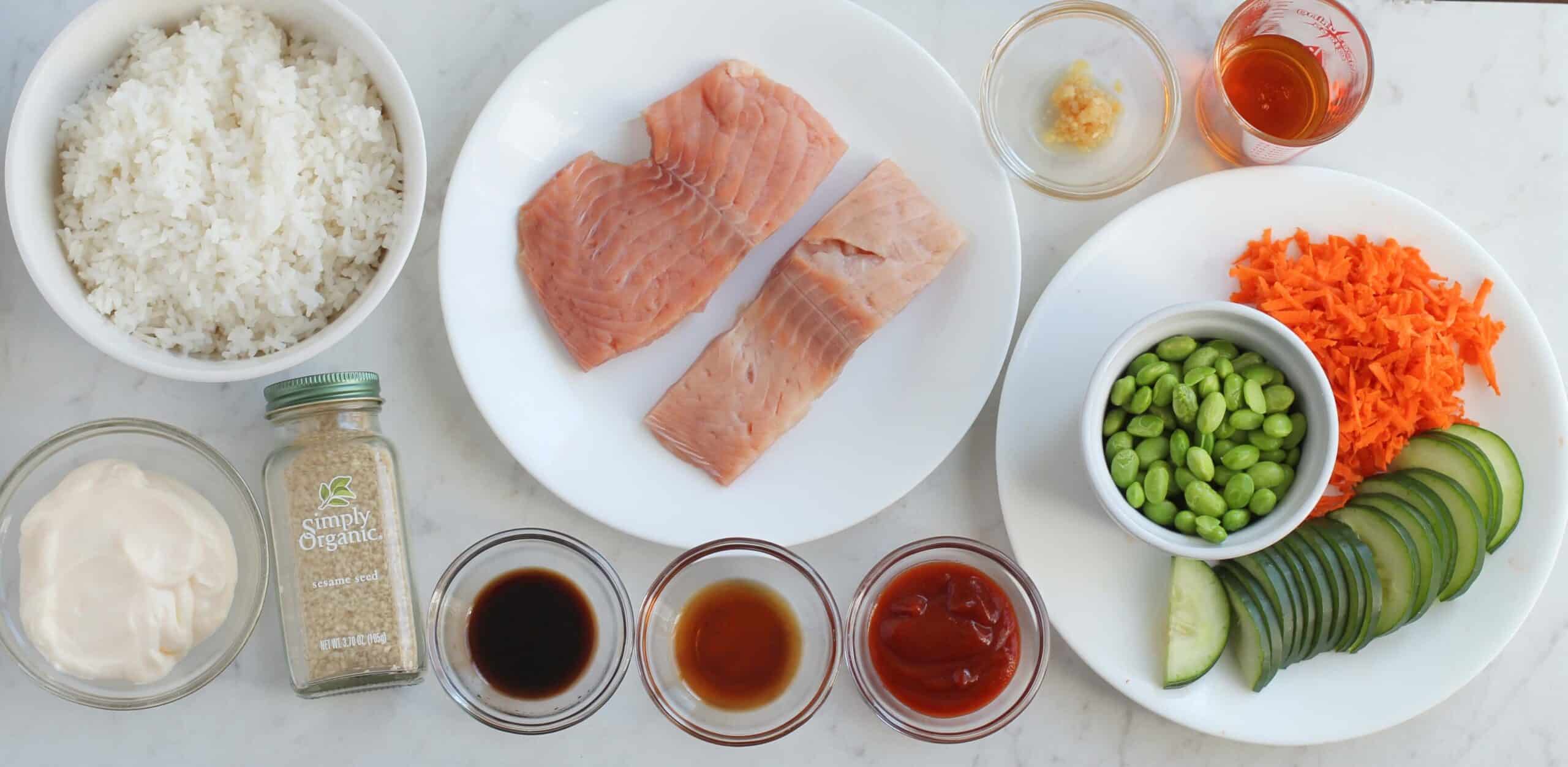 Ingredients fish include cooked rice, two raw fish fillets, grated carrots, edamame, cucumber slices, and various condiments in small bowls on a countertop.