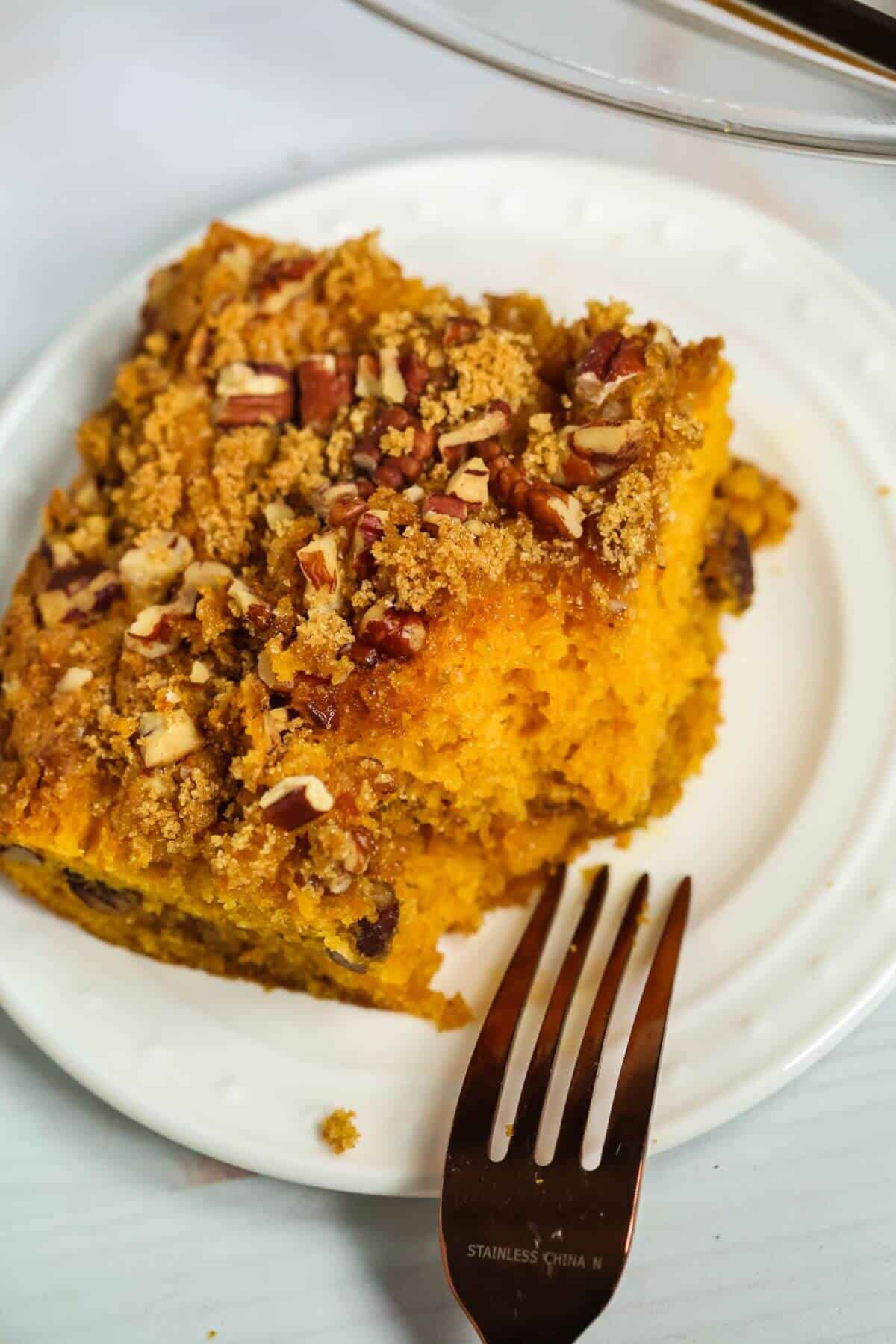 A slice of pumpkin coffee cake topped with crumbly streusel and chopped pecans sits invitingly on a white plate beside a fork, reminiscent of the richness you might find in a butterscotch cake.