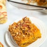 A square slice of pumpkin cake topped with pecans and streusel graces a white plate, reminiscent of delicious butterscotch cake. Nearby, a pan holds more slices, while a patterned cloth peeks into view, adding charm to the cozy scene.
