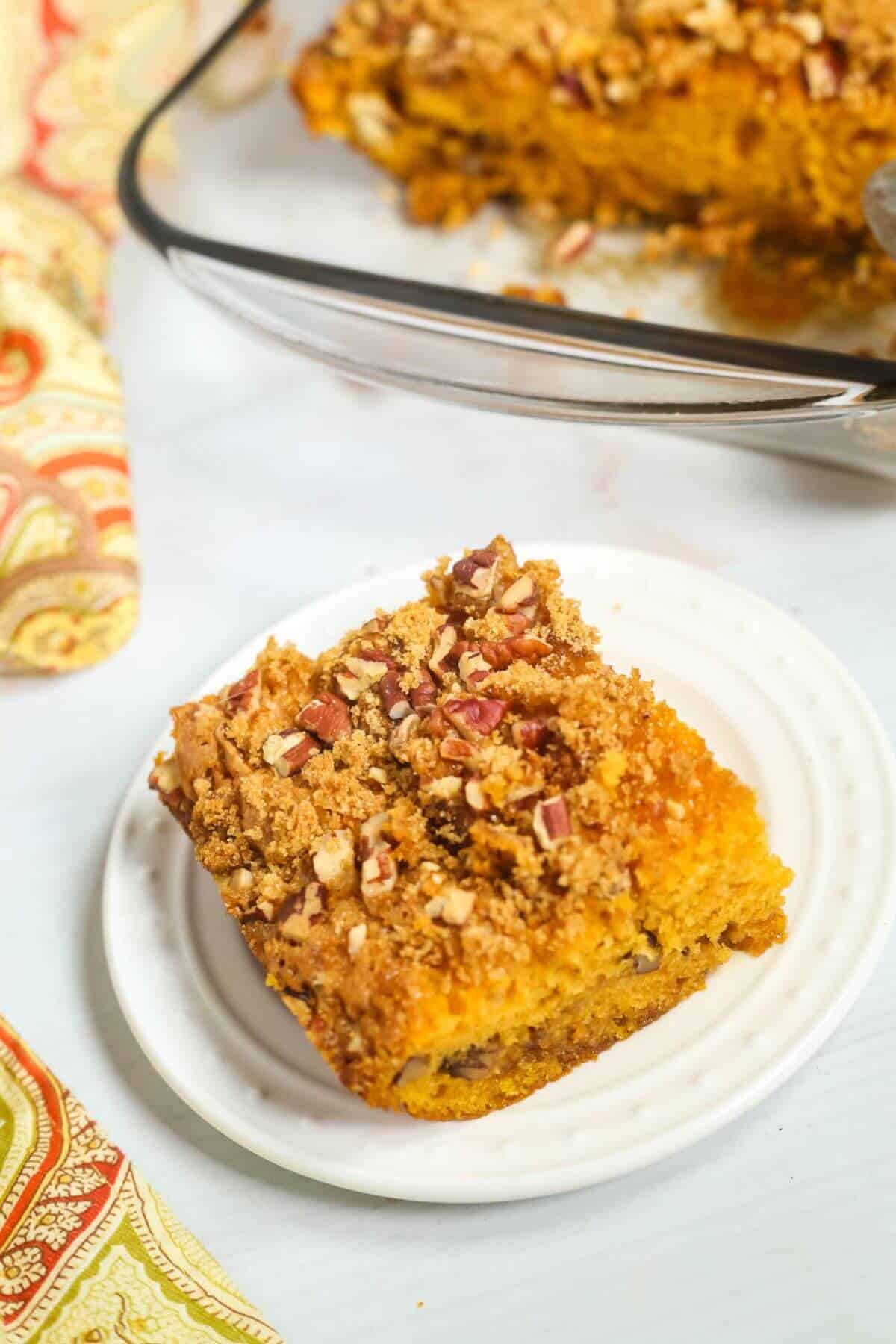 A square slice of pumpkin cake topped with pecans and streusel graces a white plate, reminiscent of delicious butterscotch cake. Nearby, a pan holds more slices, while a patterned cloth peeks into view, adding charm to the cozy scene.