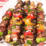 Skewers of grilled vegetables, including peppers, onions, and mushrooms, along with juicy pieces of steak kabobs are artfully arranged on a white plate.