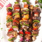 Four grilled steak kabobs and vegetable skewers on a white plate. The skewers include beef, bell peppers, mushrooms, onions, and herbs.