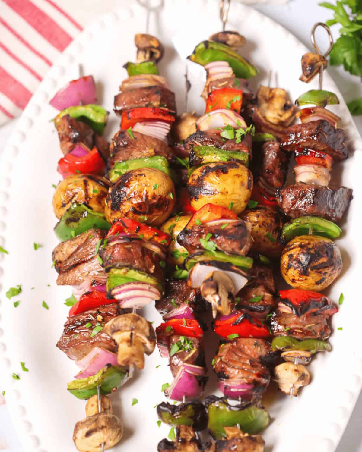 Four grilled steak kabobs and vegetable skewers on a white plate. The skewers include beef, bell peppers, mushrooms, onions, and herbs.