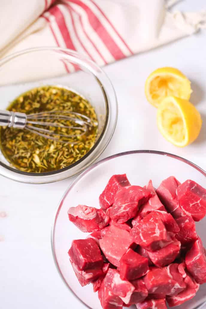 A bowl of raw meat cubes next to an herb marinade with a whisk, ready for Steak Kabobs. Two halved, squeezed lemons and a striped cloth are in the background.