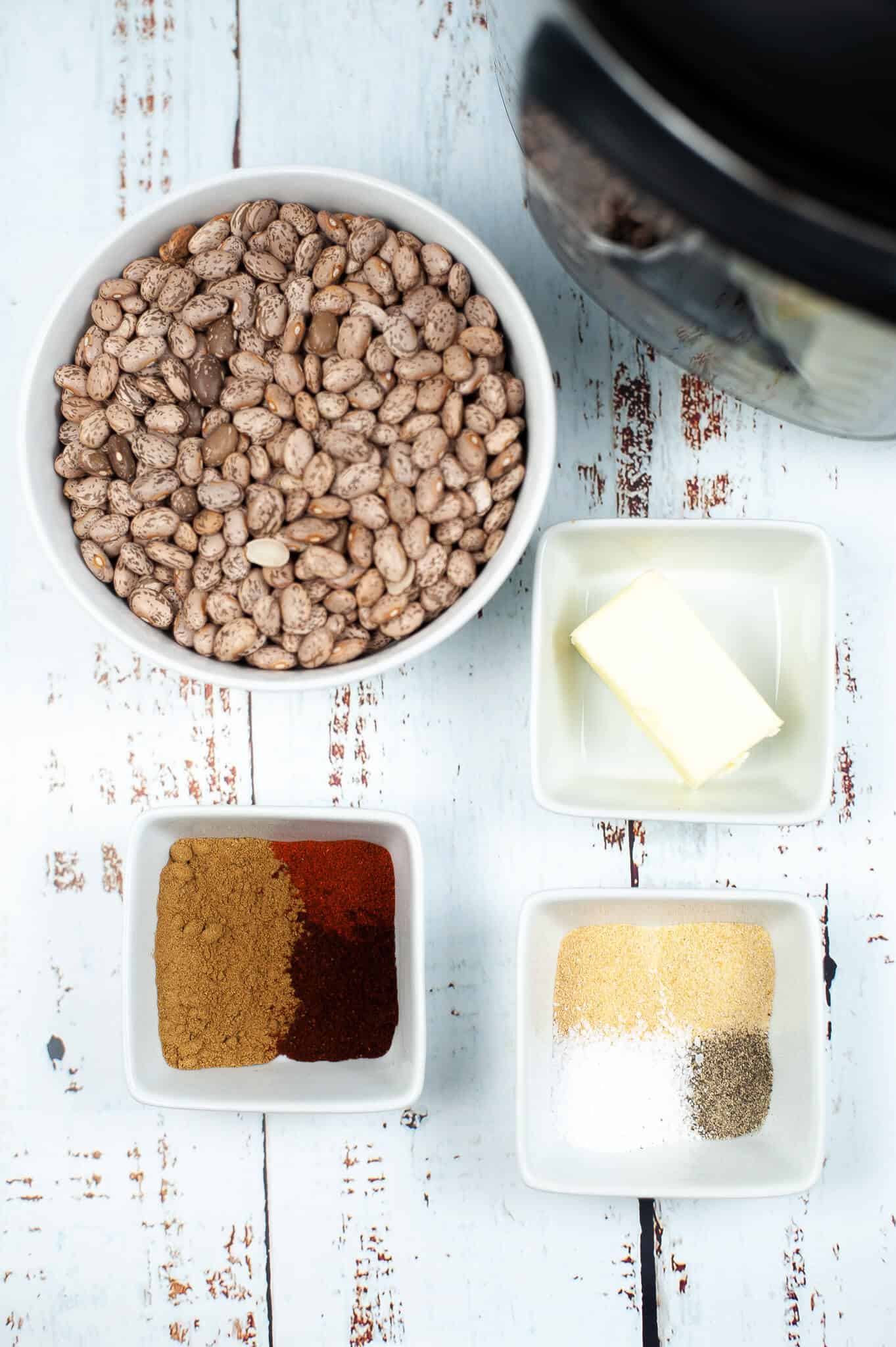 Overhead view of bowls with pinto beans, butter, and spices including chili powder, white salt, and black pepper on a rustic wooden table .