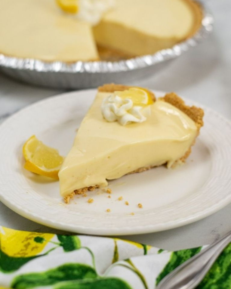 A slice of Lemon Icebox Pie with a dollop of whipped cream on a white plate, with additional lemon slices on the side.