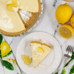 A slice of Lemon Icebox Pie served on a white plate with whole lemons and a pie in the background.
