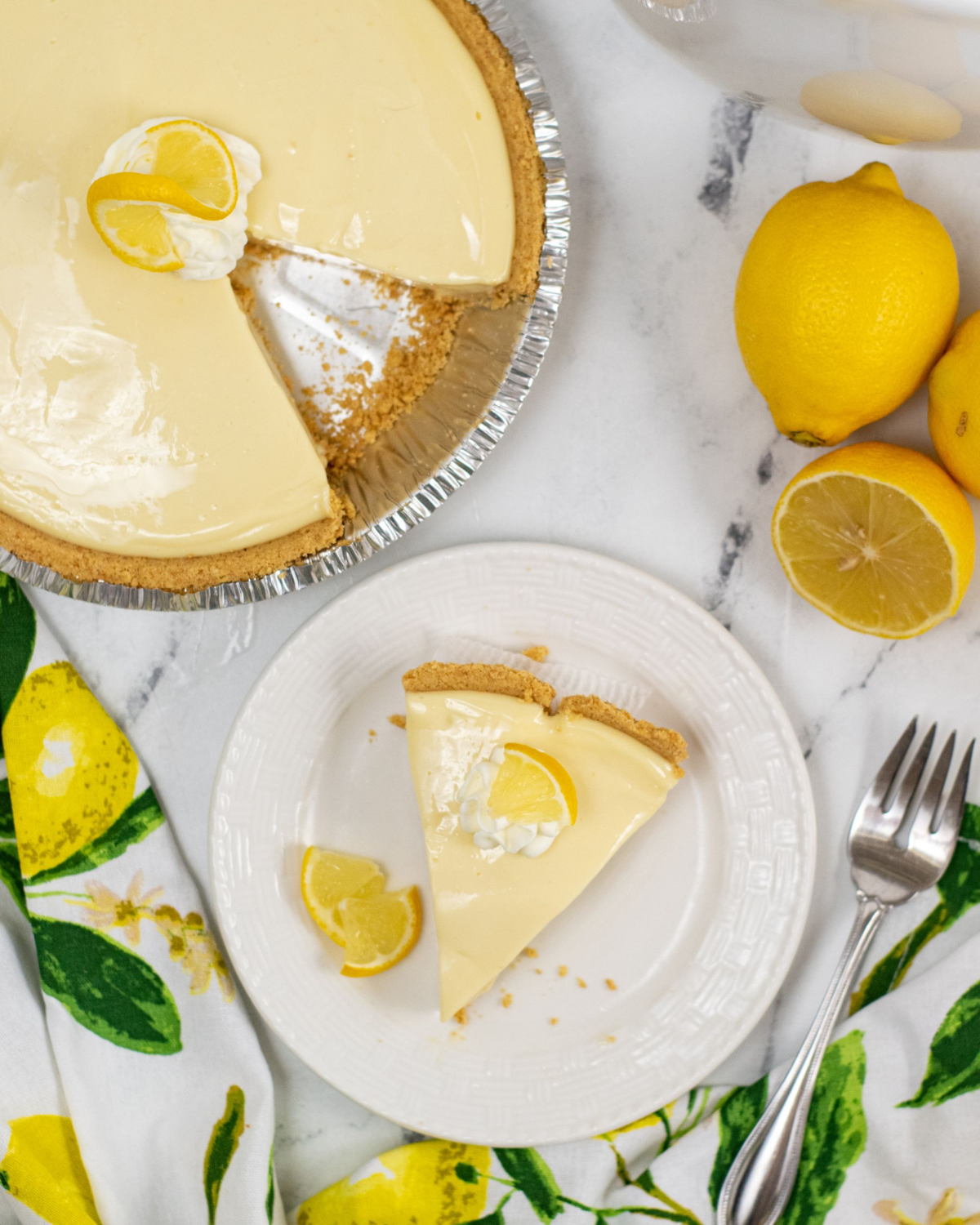 A slice of Lemon Icebox Pie served on a white plate with whole lemons and a pie in the background.