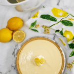 A Lemon Icebox Pie with a creamy topping and lemon slice garnish, accompanied by fresh lemons and a tea setting.