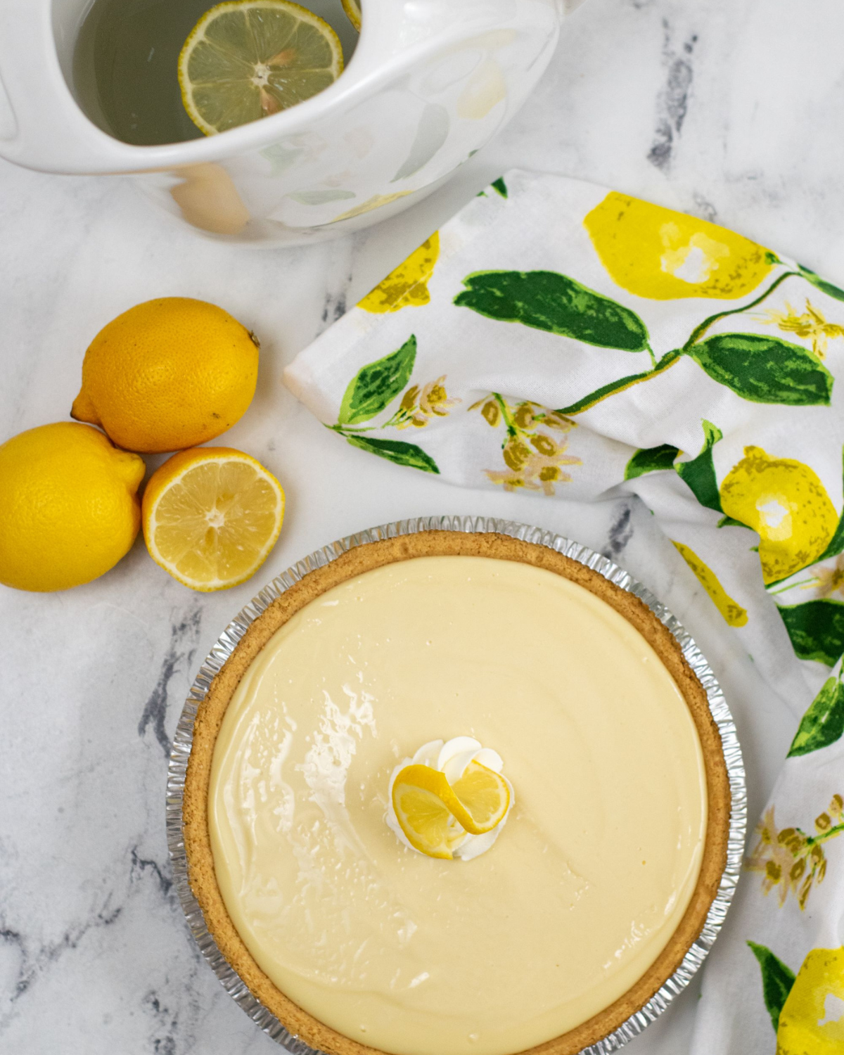 A Lemon Icebox Pie with a creamy topping and lemon slice garnish, accompanied by fresh lemons and a tea setting.