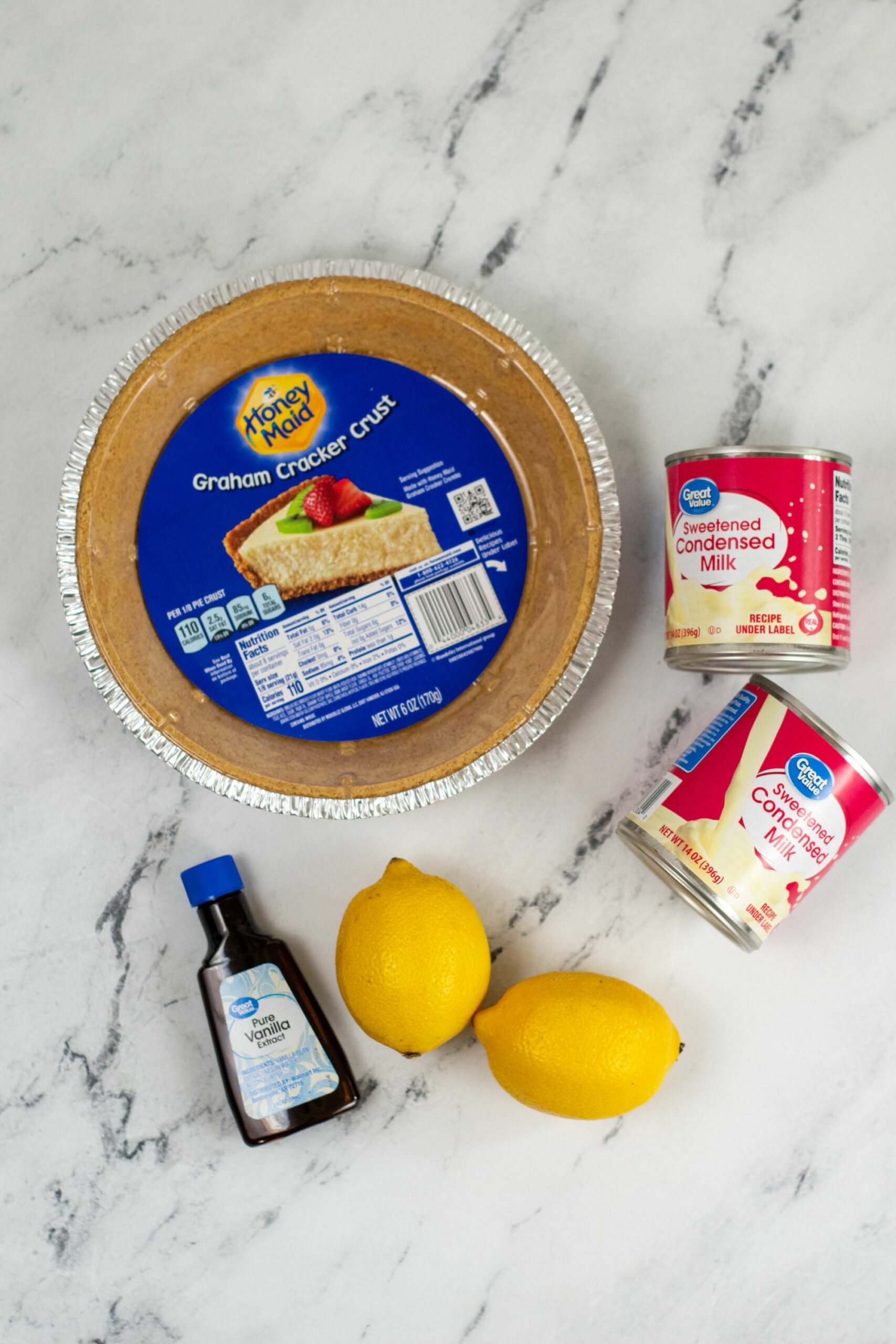 Ingredients for Lemon Icebox Pie on a marble surface, including a graham cracker crust, condensed milk, lemons, and vanilla extract.