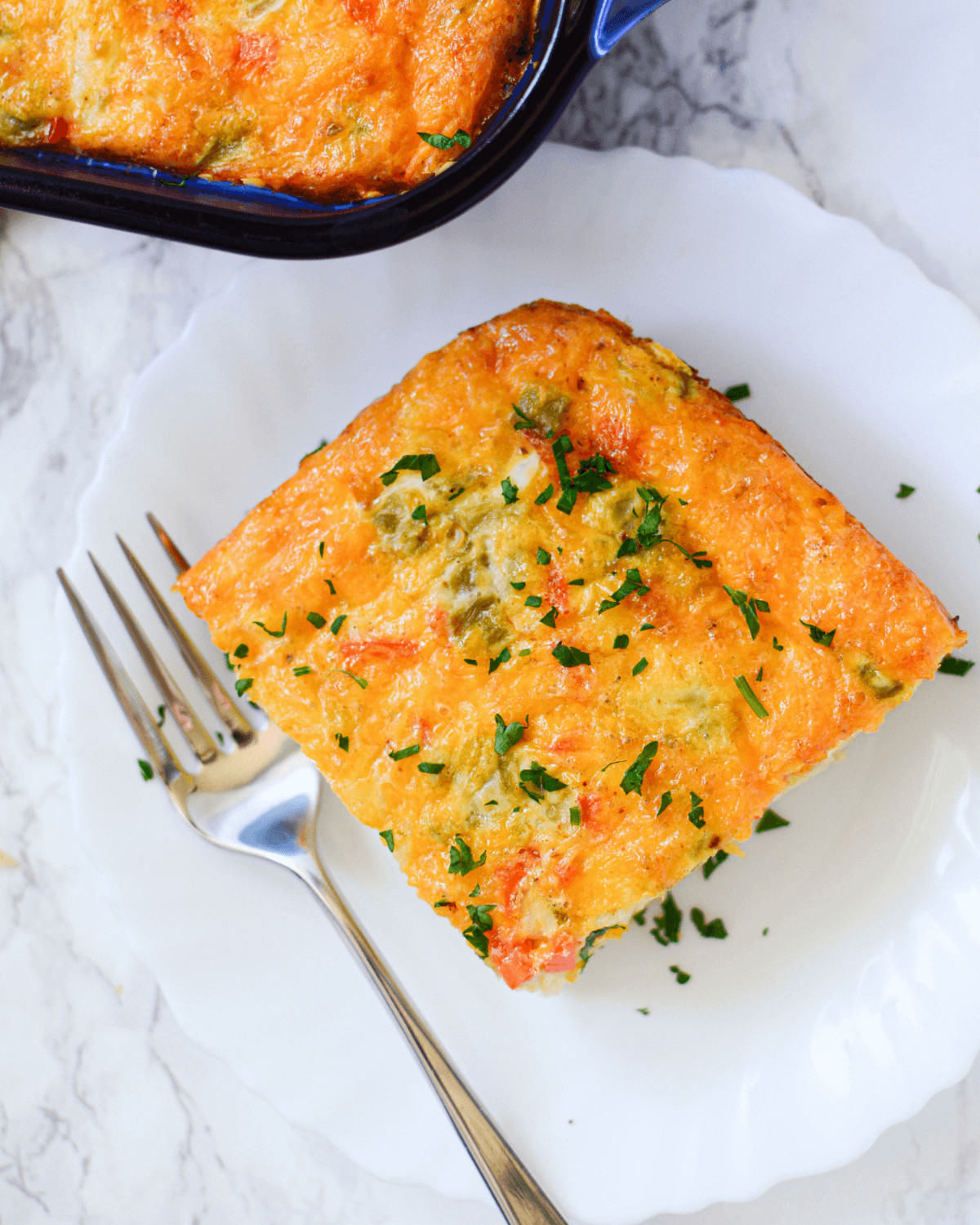 A square slice of cheesy Mexican breakfast casserole on a white plate with a fork, garnished with chopped parsley.