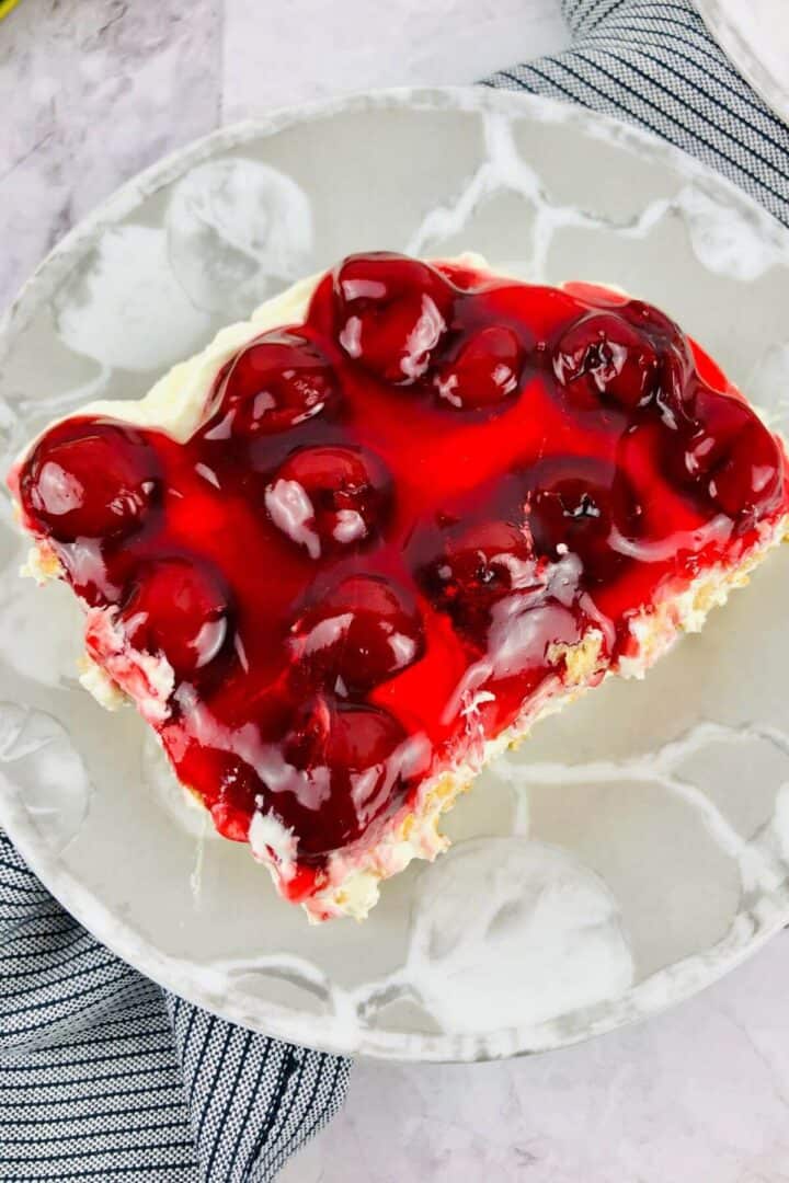 A slice of No Bake Cherry Cheesecake with a creamy layer sits on a white marbled plate, topped with cherries and placed next to a blue-striped cloth.