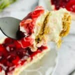 A fork holding a bite of luscious No Bake Cherry Cheesecake with graham cracker crust, with more creamy cheesecake and cherry topping visible on a plate in the background.