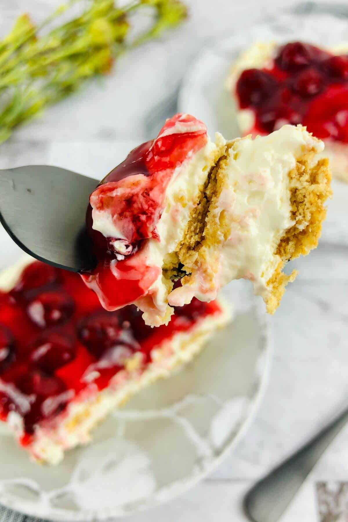 A fork holding a bite of luscious No Bake Cherry Cheesecake with graham cracker crust, with more creamy cheesecake and cherry topping visible on a plate in the background.