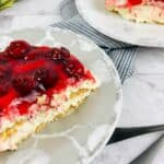 Two plates with slices of No Bake Cherry Cheesecake, featuring a creamy layer and crumbly base, set on a marble surface with utensils and flowers in the background.