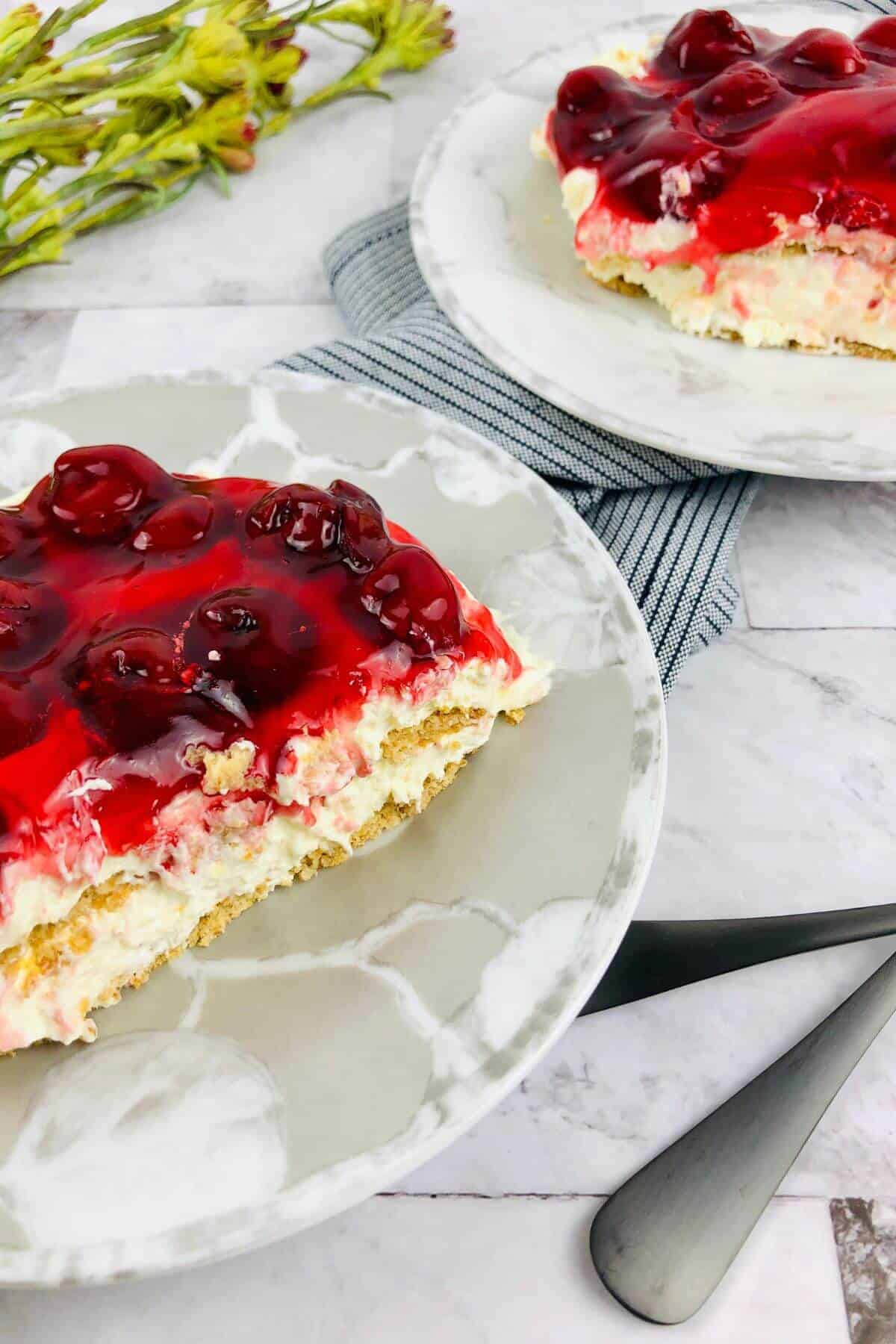 Two plates with slices of No Bake Cherry Cheesecake, featuring a creamy layer and crumbly base, set on a marble surface with utensils and flowers in the background.