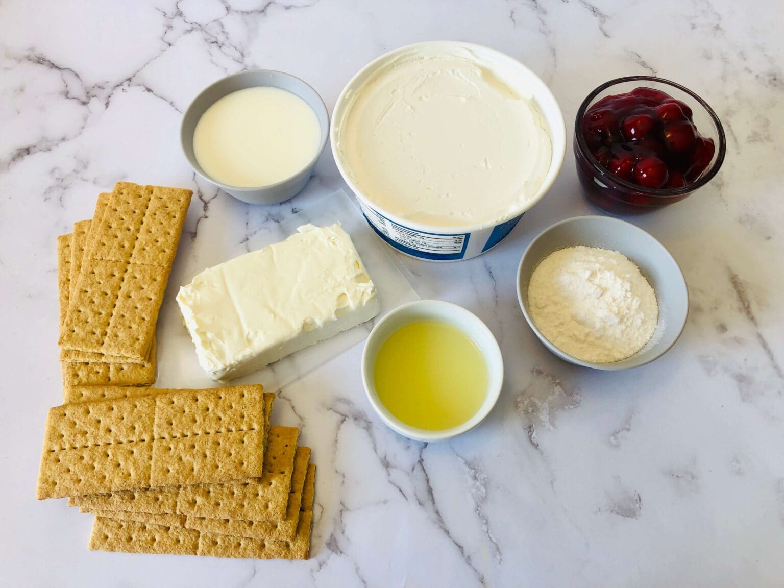Seven ingredients on a marble surface: graham crackers, cream cheese, whipped topping, cherries, heavy cream, powdered sugar, and melted butter—everything you need for a classic dessert.