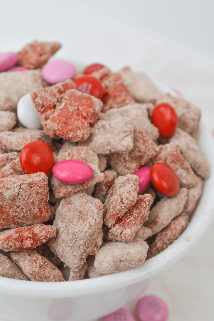 A bowl of Chex muddy recipe delights, featuring cinnamon-sugar coated snack pieces mixed with red, pink, and white candy-coated chocolates.