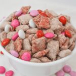 A white bowl filled with chocolate-covered cereal pieces, reminiscent of a classic Chex muddy recipe, dusted in powdered sugar and adorned with red, pink, and white candy-coated chocolates.