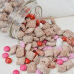 A jar tipped over, revealing cereal squares dusted in powdered sugar and sprinkled with pink, red, and white candy-coated chocolates onto a pristine white surface.