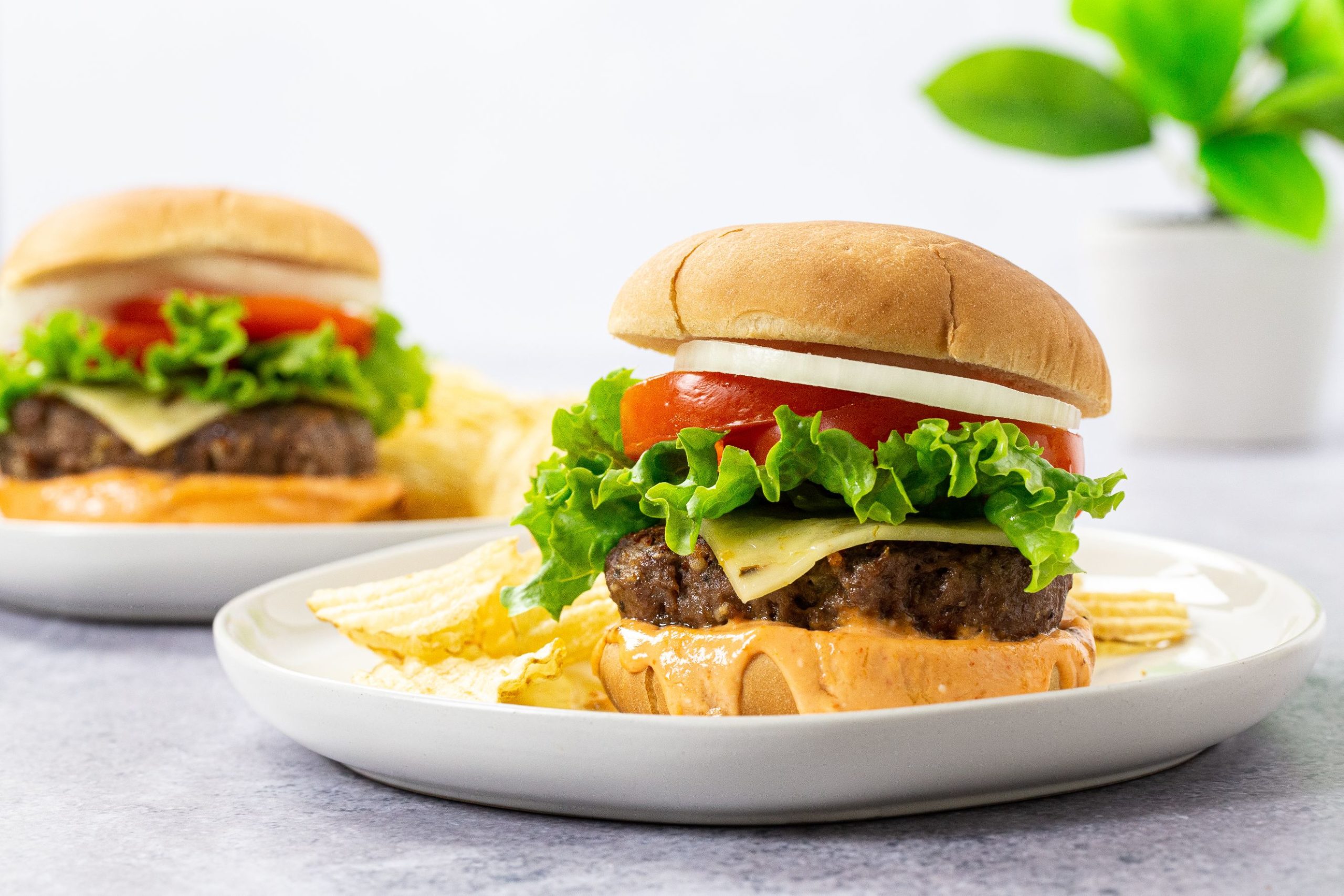 Two spicy burgers on white plates with cheese lettuce and tomatoes.
