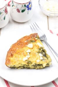A slice of spinach and feta quiche sits invitingly on a white plate with a fork, set against a pristine background adorned with decorative ceramic containers. Similar to a classic spinach frittata recipe, every bite promises delightful flavors.
