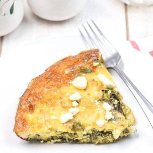 A slice of spinach and feta quiche sits invitingly on a white plate with a fork, set against a pristine background adorned with decorative ceramic containers. Similar to a classic spinach frittata recipe, every bite promises delightful flavors.