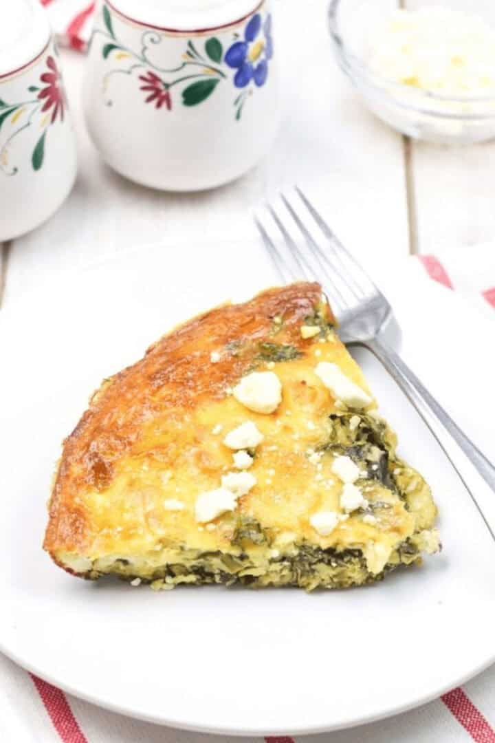 A slice of spinach and feta quiche sits invitingly on a white plate with a fork, set against a pristine background adorned with decorative ceramic containers. Similar to a classic spinach frittata recipe, every bite promises delightful flavors.