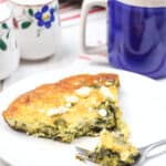 A slice of spinach and cheese frittata rests on a white plate with a fork, perfectly paired with a blue mug of coffee. Floral-patterned shakers sit elegantly on a striped cloth, bringing the delicious Spinach Frittata recipe to life in this cozy setting.