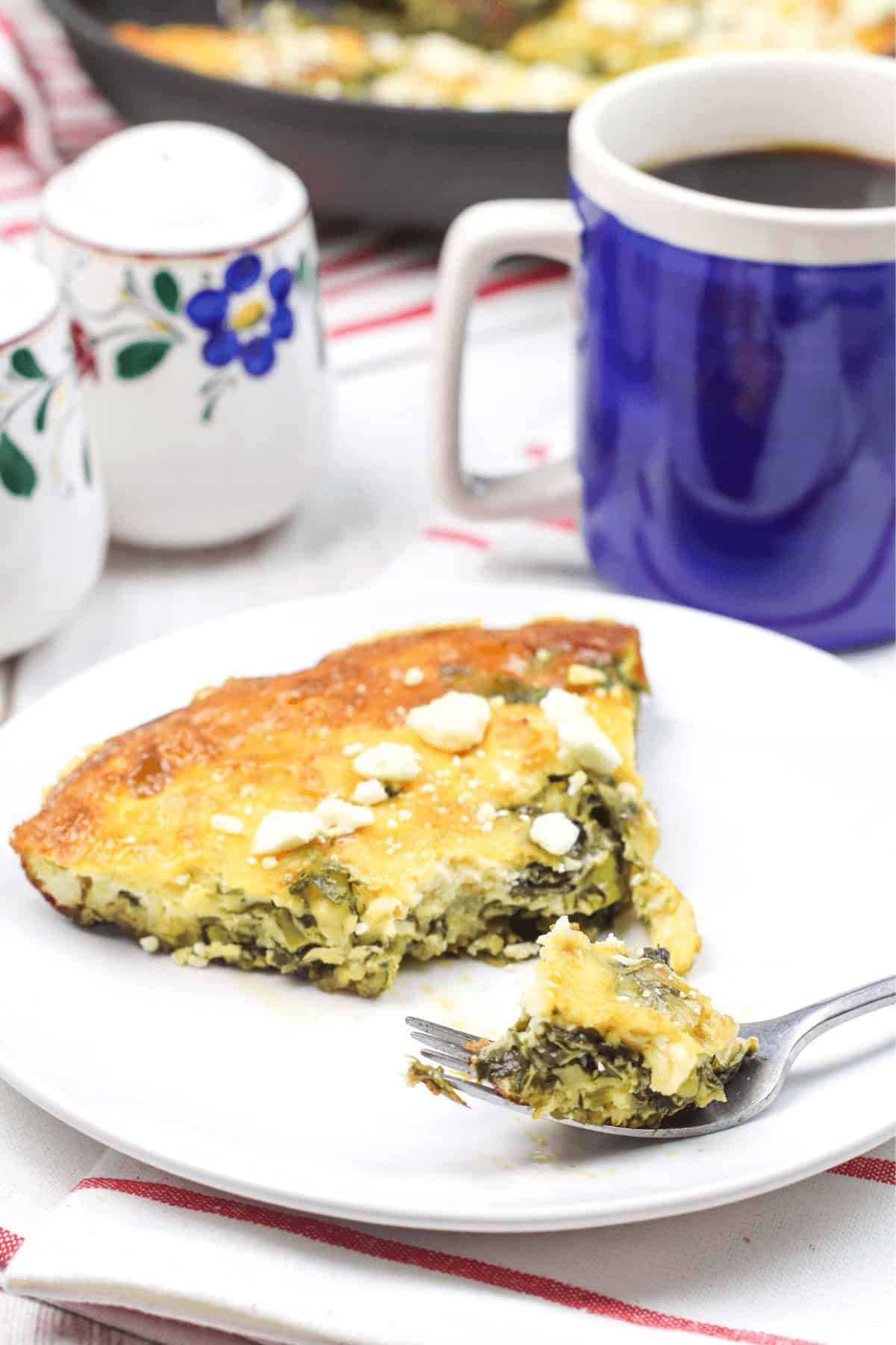 A slice of spinach and cheese frittata rests on a white plate with a fork, perfectly paired with a blue mug of coffee. Floral-patterned shakers sit elegantly on a striped cloth, bringing the delicious Spinach Frittata recipe to life in this cozy setting.