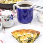 A slice of delicious breakfast pie sits on a white plate, next to a blue mug of coffee and a sugar bowl. The skillet brimming with more of this delightful spinach frittata recipe rests on a red striped cloth.