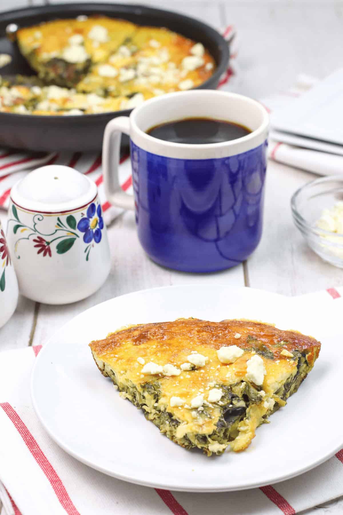 A slice of delicious breakfast pie sits on a white plate, next to a blue mug of coffee and a sugar bowl. The skillet brimming with more of this delightful spinach frittata recipe rests on a red striped cloth.