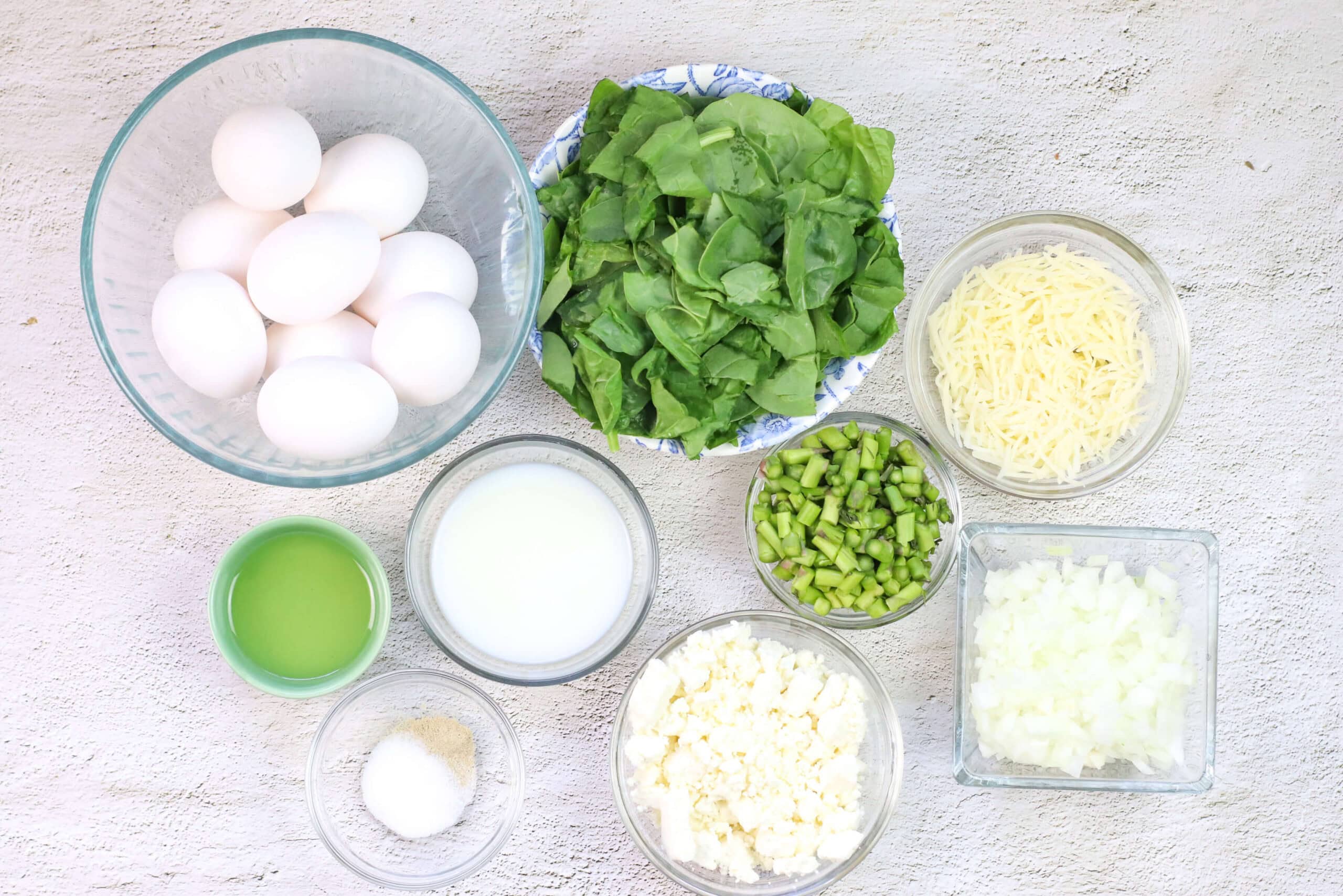Bowls of eggs, fresh spinach, grated cheese, chopped green onions, milk, feta, salt, and a verdant green liquid artfully arranged on a textured surface. A vibrant tableau for crafting your favorite Spinach Frittata recipe.