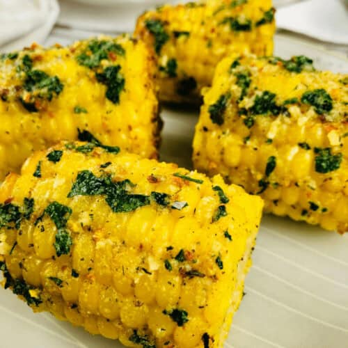 Close-up of air fryer corn on the cob pieces, garnished with chopped herbs and spices, arranged on a white plate.