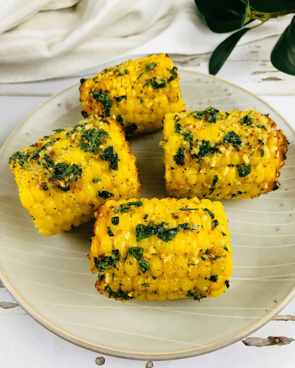 Four pieces of corn on the cob in air fryer seasoned with herbs and spices, are arranged on a light-colored plate. A white cloth and green leaves are visible in the background.