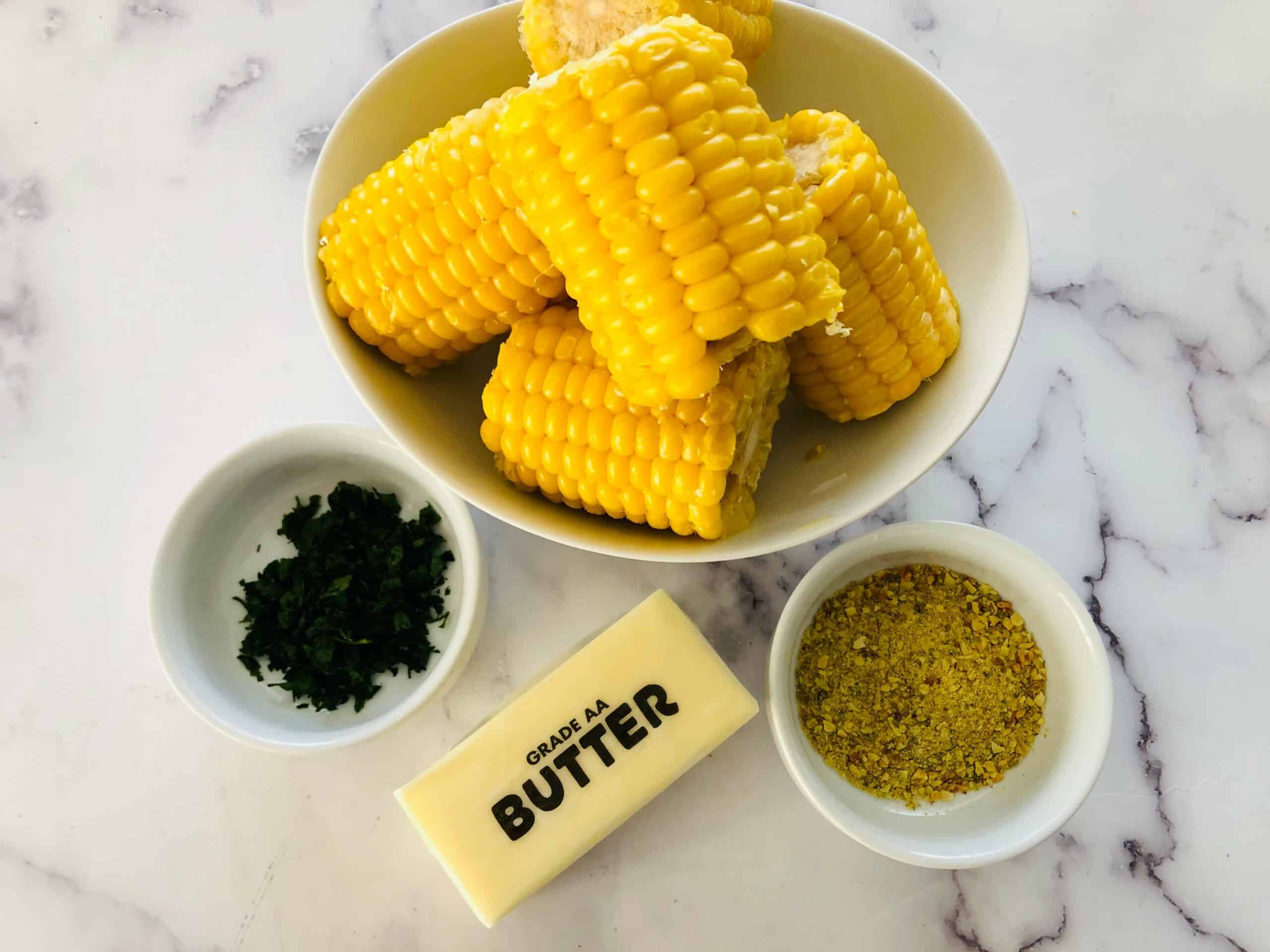 A bowl of corn on the cob, a stick of butter, a small dish of chopped herbs, and a small bowl of yellow seasoning are arranged on a marble countertop, ready to be transformed into delicious Air Fryer Corn.