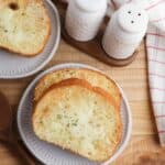 Two slices of cheesy toast are served on a plate, accompanied by salt and pepper shakers and a wooden spoon on a rustic wooden table.