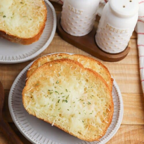 Two slices of Air Fryer Garlic Bread with melted cheese and herbs rest on a plate, with salt and pepper shakers in the background on a wooden table.