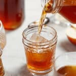 A hand pours apple pie moonshine from a glass jar into a glass filled with ice cubes on a white surface, with additional jars and a sliced apple in the background, evoking the cozy feeling of enjoying apple pie moonshine.