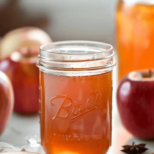 A glass jar filled with apple pie moonshine, surrounded by fresh apples and spices, sits on a countertop.