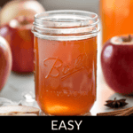 A jar of apple pie moonshine sits on a table with fresh apples, a cinnamon stick, and star anise, with the text "Easy Apple Pie Moonshine" elegantly displayed below.
