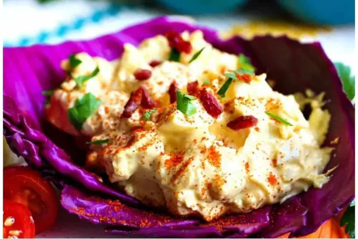 Discover a delightfully easy egg salad recipe: Egg salad topped with crispy bacon bits and fresh herbs, elegantly served in a purple cabbage leaf and finished with a sprinkle of paprika.