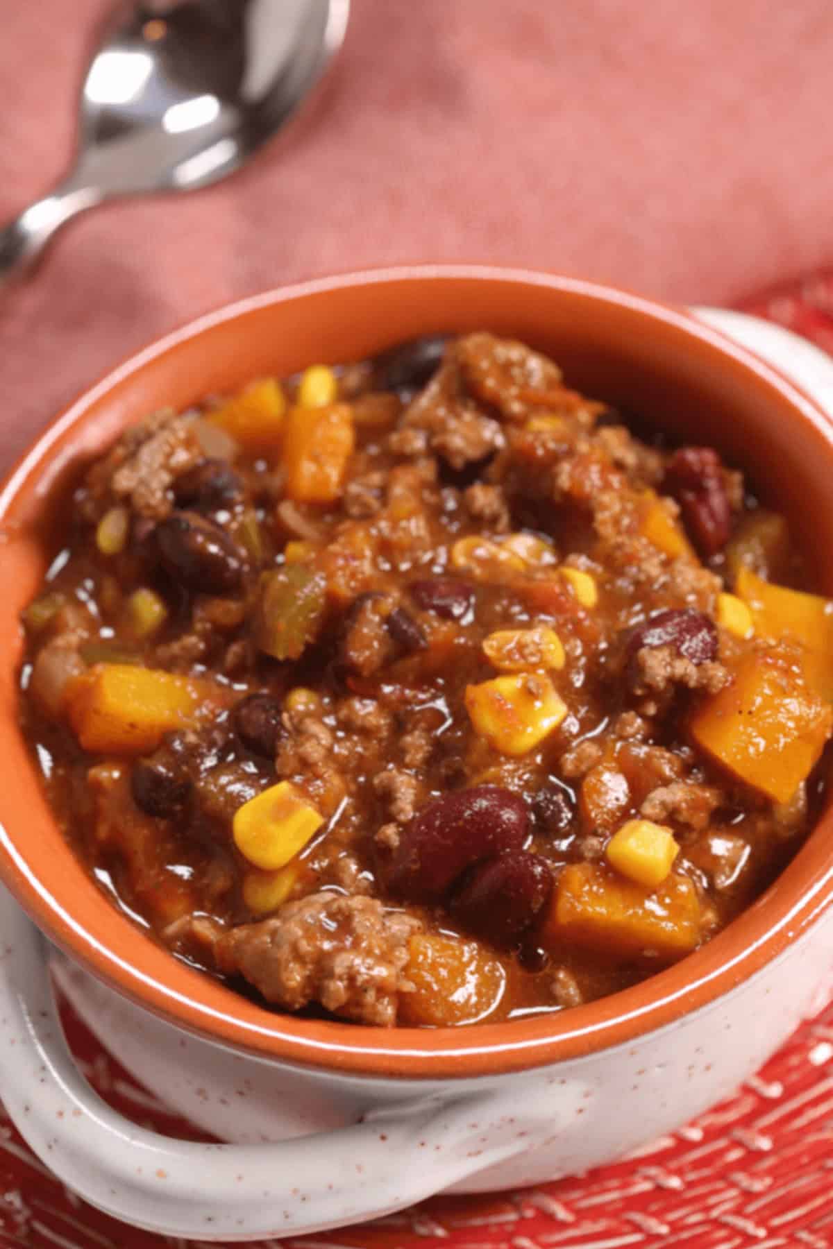 A bowl of beef and bean chili with corn, diced vegetables, and a rich tomato-based sauce, placed on a red and white mat, with a spoon in the background.