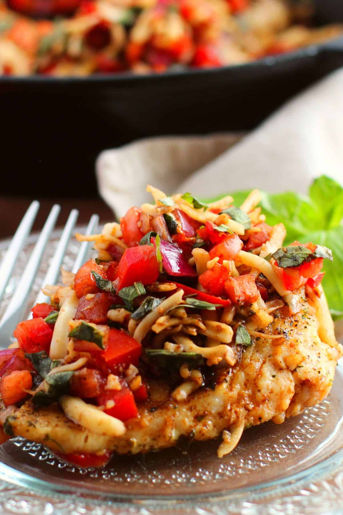 Grilled chicken breast topped with a savory bruschetta chicken recipe of diced tomatoes, red onions, fresh basil, and shredded cheese on a clear glass plate with a fork.