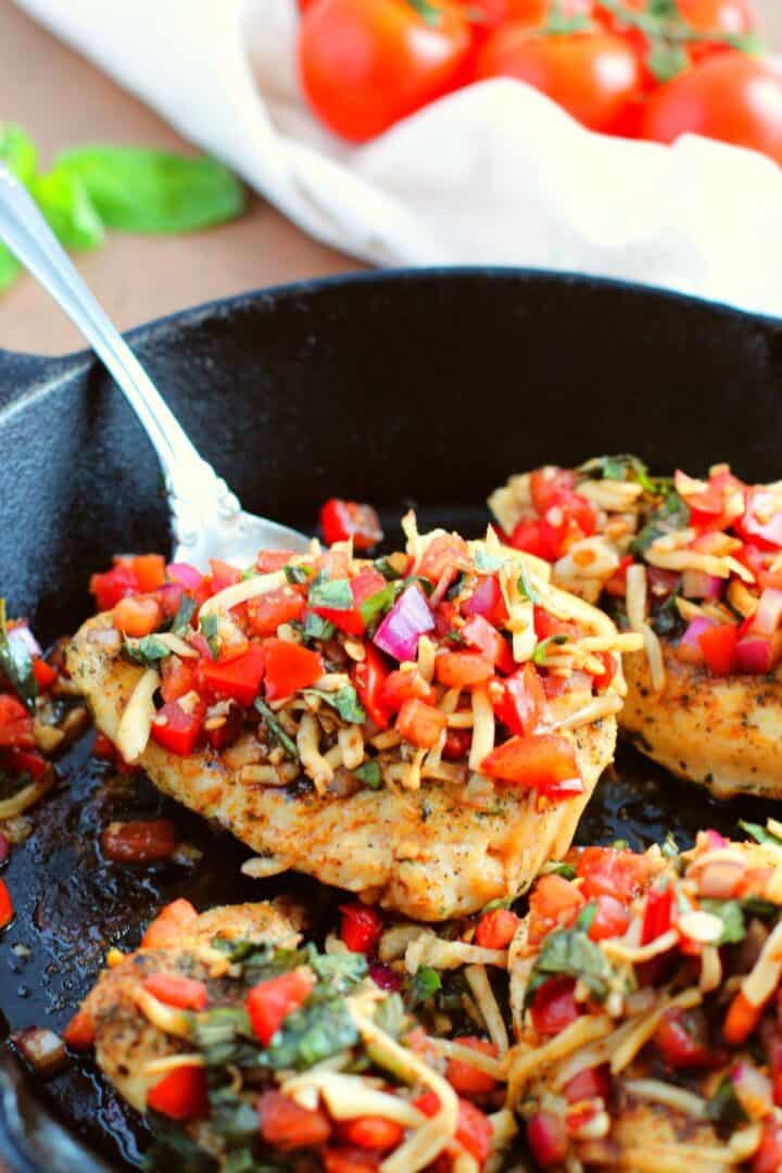 Seasoned meat in a cast iron skillet, topped with chopped tomatoes, red onion, basil, and cheese—this recipe is served with a spoon beside it. Fresh tomatoes are visible in the background.